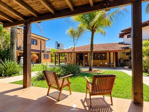 A covered patio area with two wooden chairs, a brick building, and a palm tree in the background. - Pousada Baleia Franca - Ferrugem/sc (Garopaba)