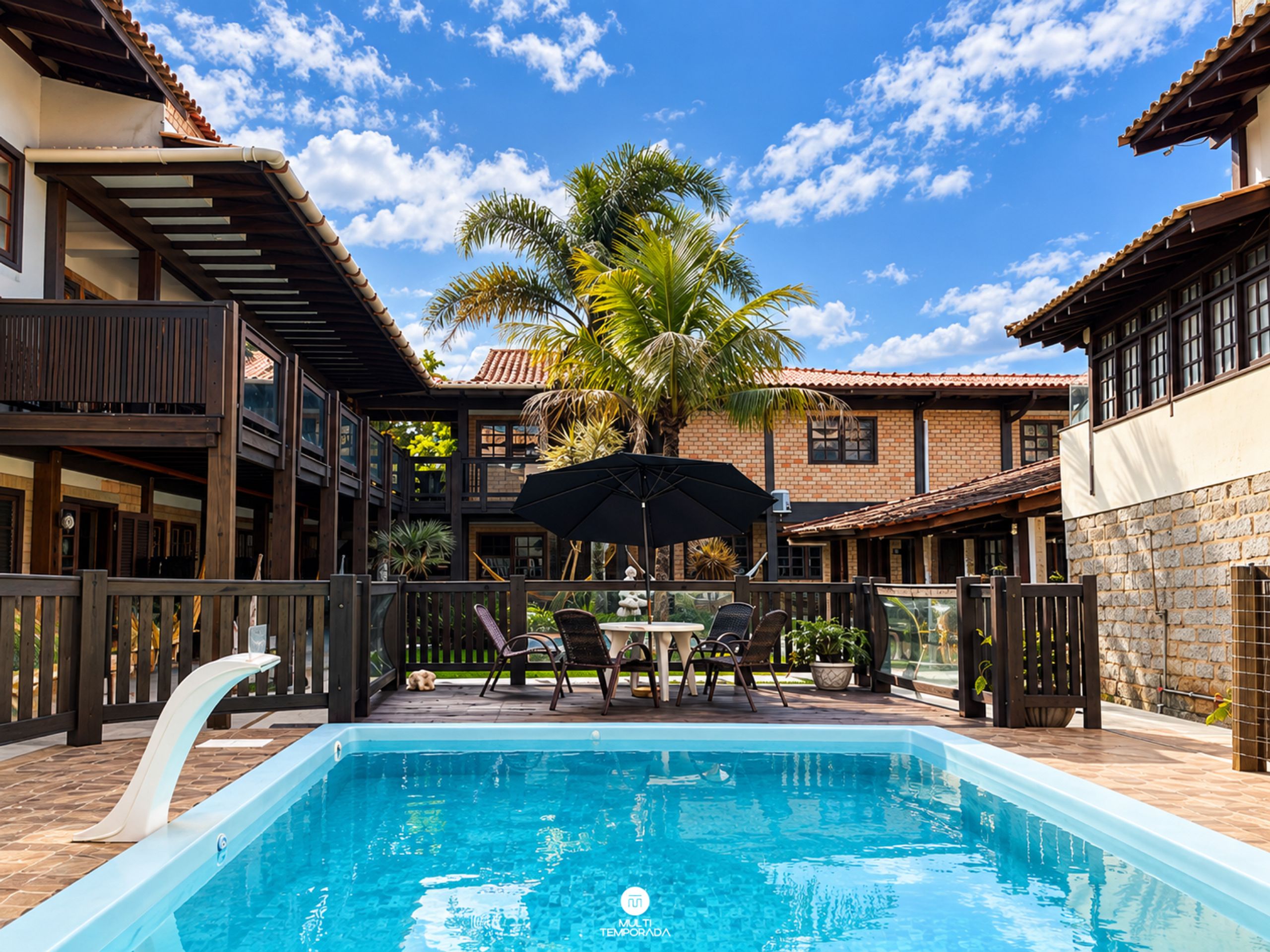 A swimming pool with a wooden deck, outdoor furniture, and a palm tree in the background.