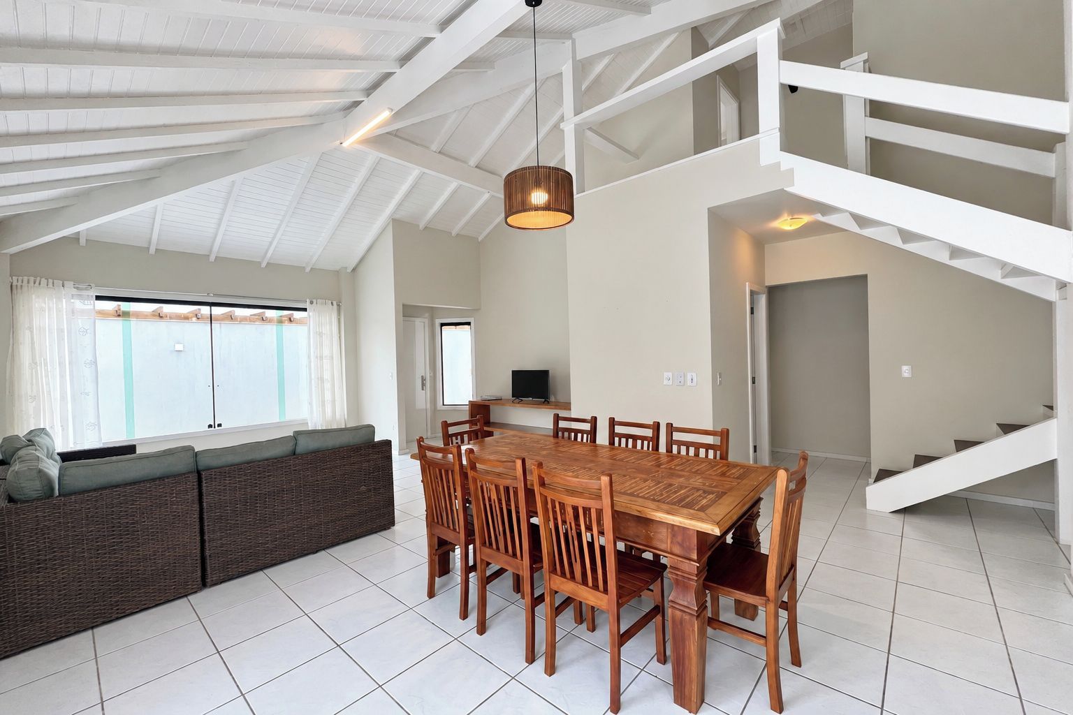 A spacious dining area with a wooden table and chairs, a high ceiling with exposed beams, and a staircase leading to an upper level.