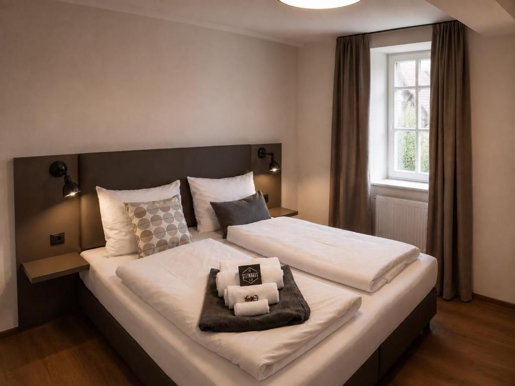 Un lit double avec du linge de lit blanc, deux tables de chevet avec des lampes, une fenêtre avec des rideaux et une petite table avec des serviettes et un panneau.