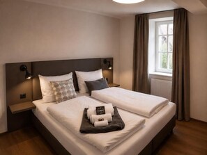A double bed with white linens, two bedside tables with lamps, a window with curtains, and a small table with towels and a sign.