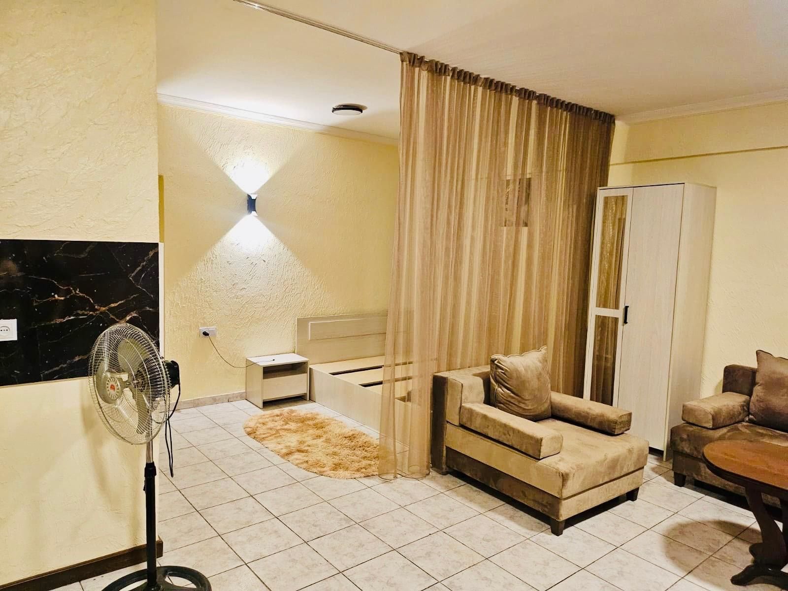 Een woonkamer met een bank, een stoel, een bijzettafeltje, een ventilator, een lamp en een garderobe.