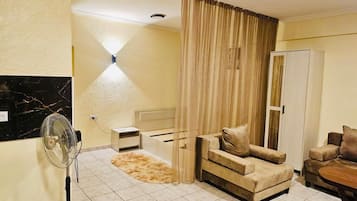 Een woonkamer met een bank, een stoel, een bijzettafeltje, een ventilator, een lamp en een garderobe.