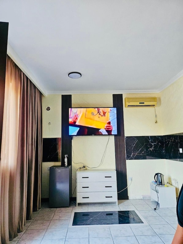 A room with a flat-screen TV mounted on the wall, a white cabinet with drawers, a small pedestal, and a tiled floor.