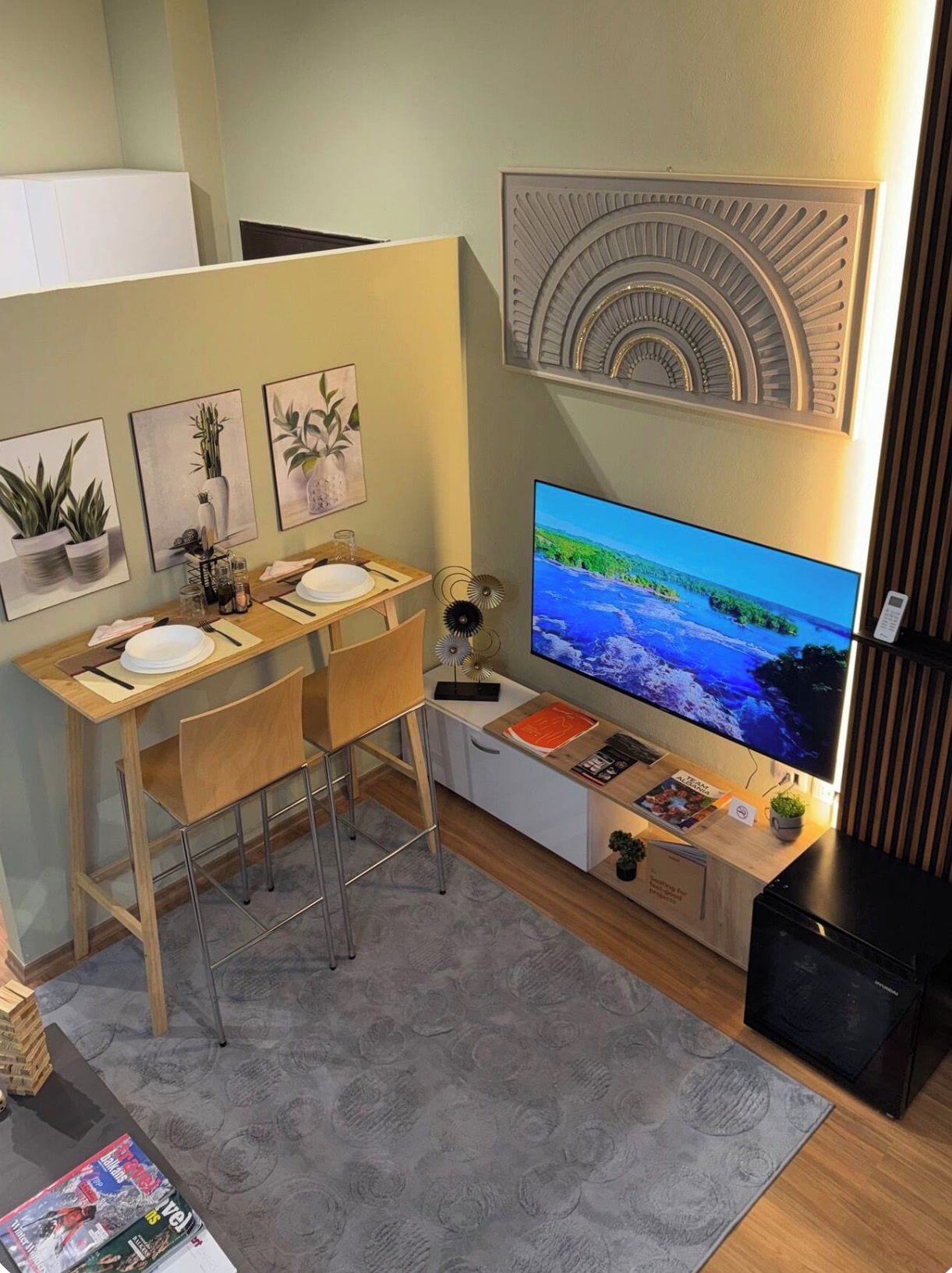 Un espace de vie compact comprenant un bureau en bois, deux chaises, un téléviseur à écran plat et un petit coin cuisine.