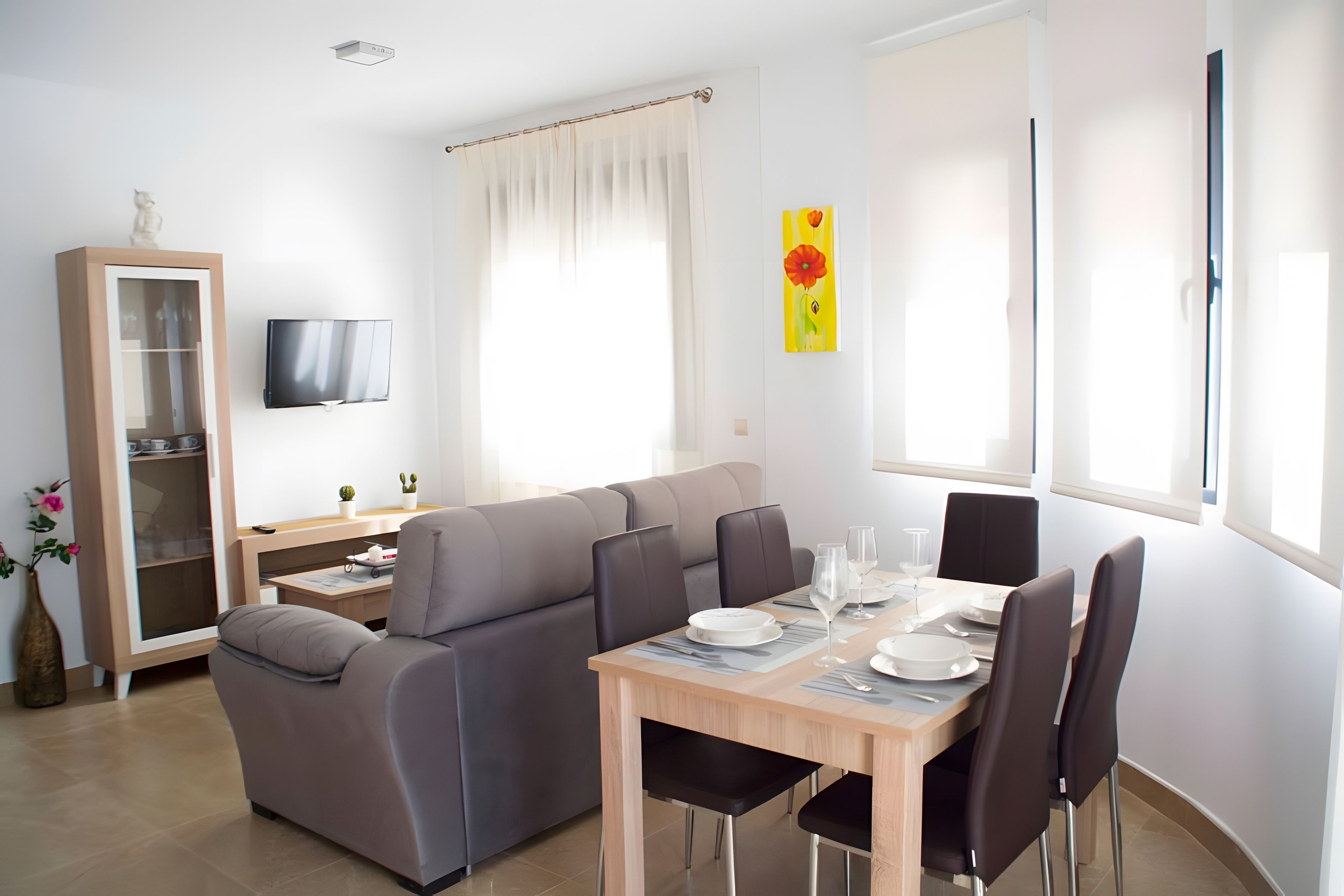 Een moderne woonkamer met een bank, een eettafel voor twee personen, een aan de muur bevestigde televisie en een kast met glazen deuren.