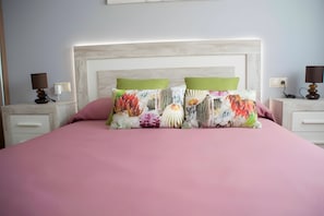 Een bed met een roze sprei, twee kussens met bloemmotieven, een wit hoofdbord en nachtlampjes op nachtkastjes.