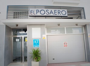 Een gebouw met een bord met de tekst 'ELPOSAERO' boven de ingang, een glazen deur, een blauw 'AT'-bord en een rookvrij-bord op de garagedeur.
