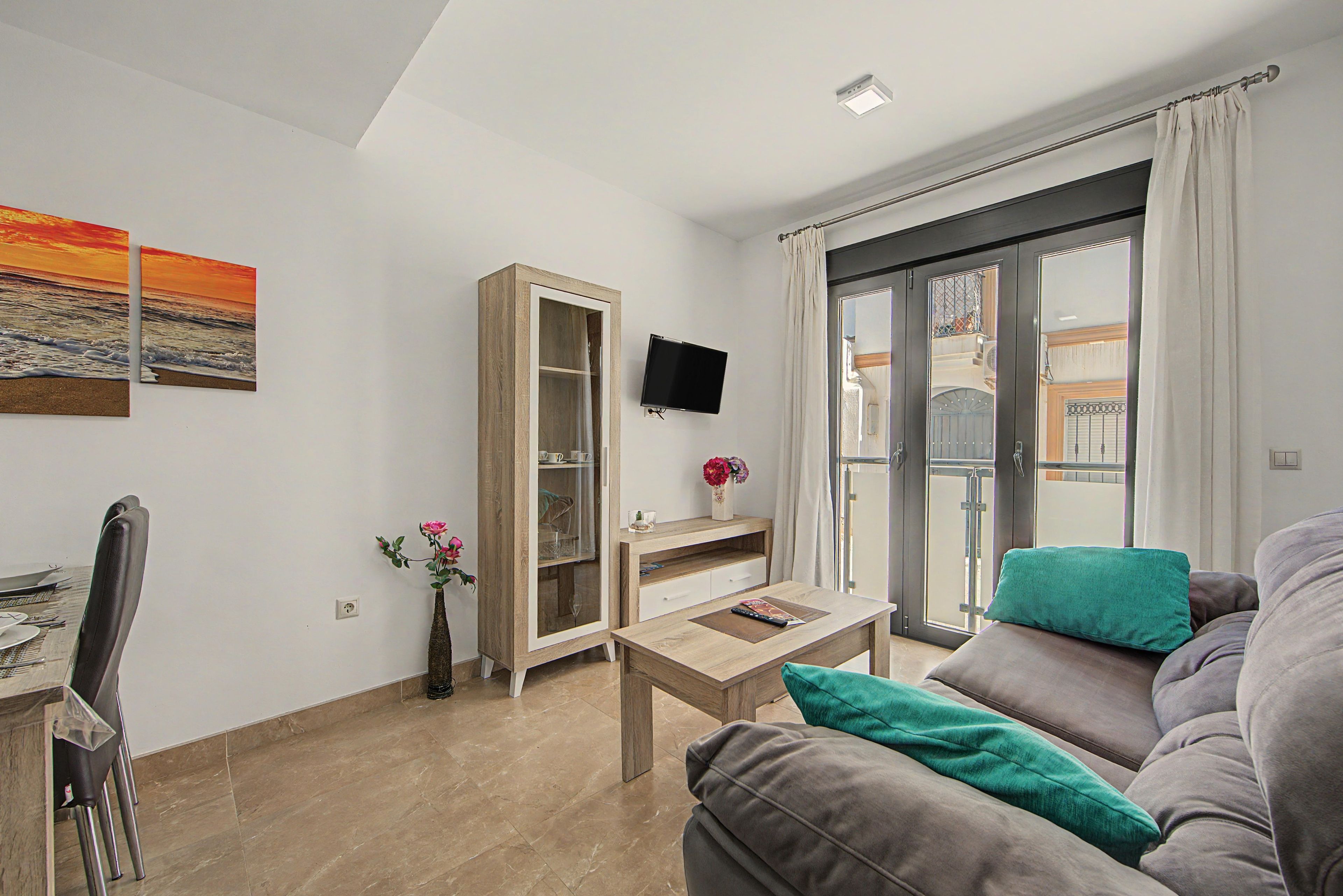 A living room with a sofa, a coffee table, a TV mounted on the wall, and a dining area with chairs and a table.