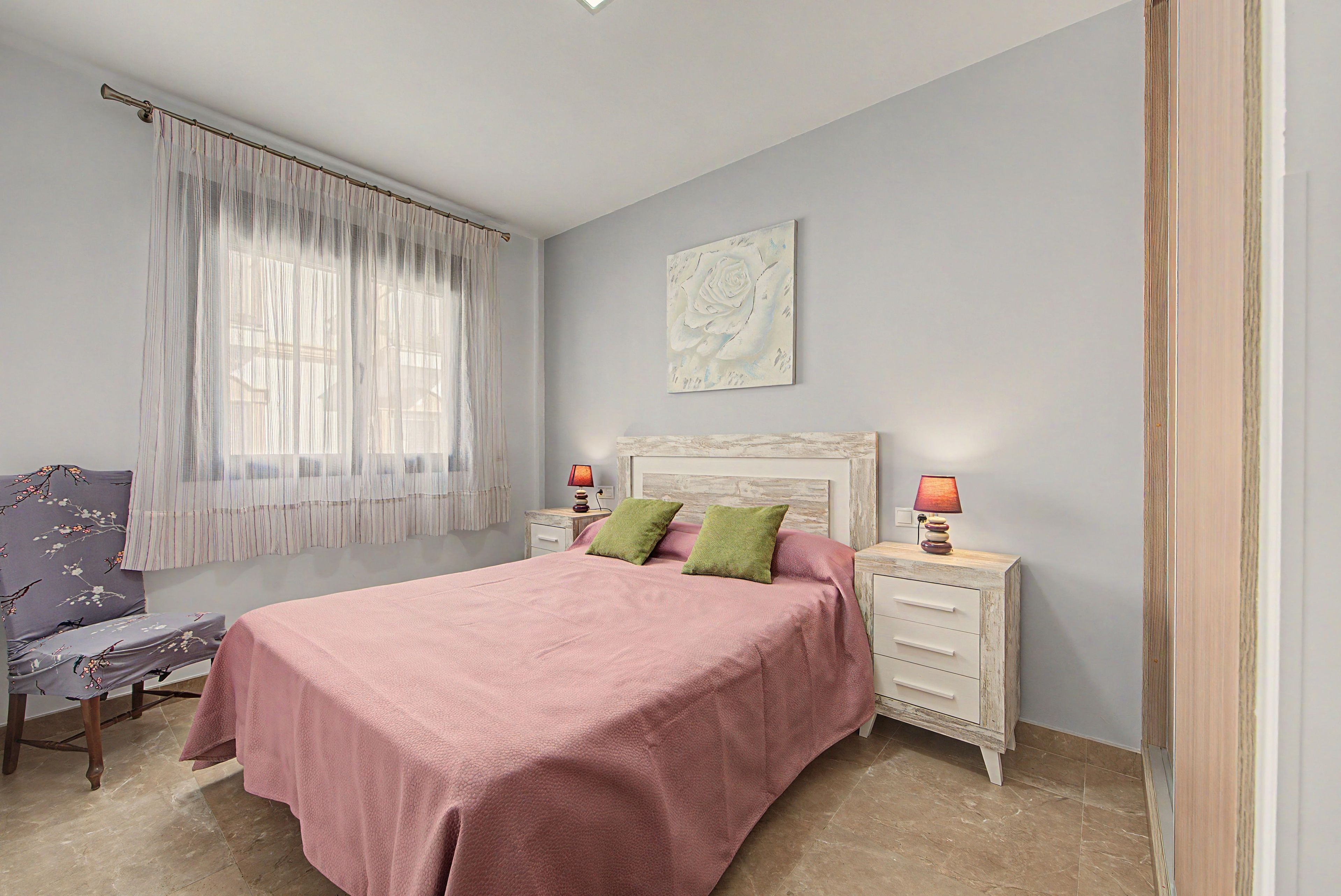 Ein Schlafzimmer mit einem Bett, zwei Nachttischen, einem Beistelltisch, einem Sessel, einem Fenster mit leichtem Vorhang und einem Wandbild.