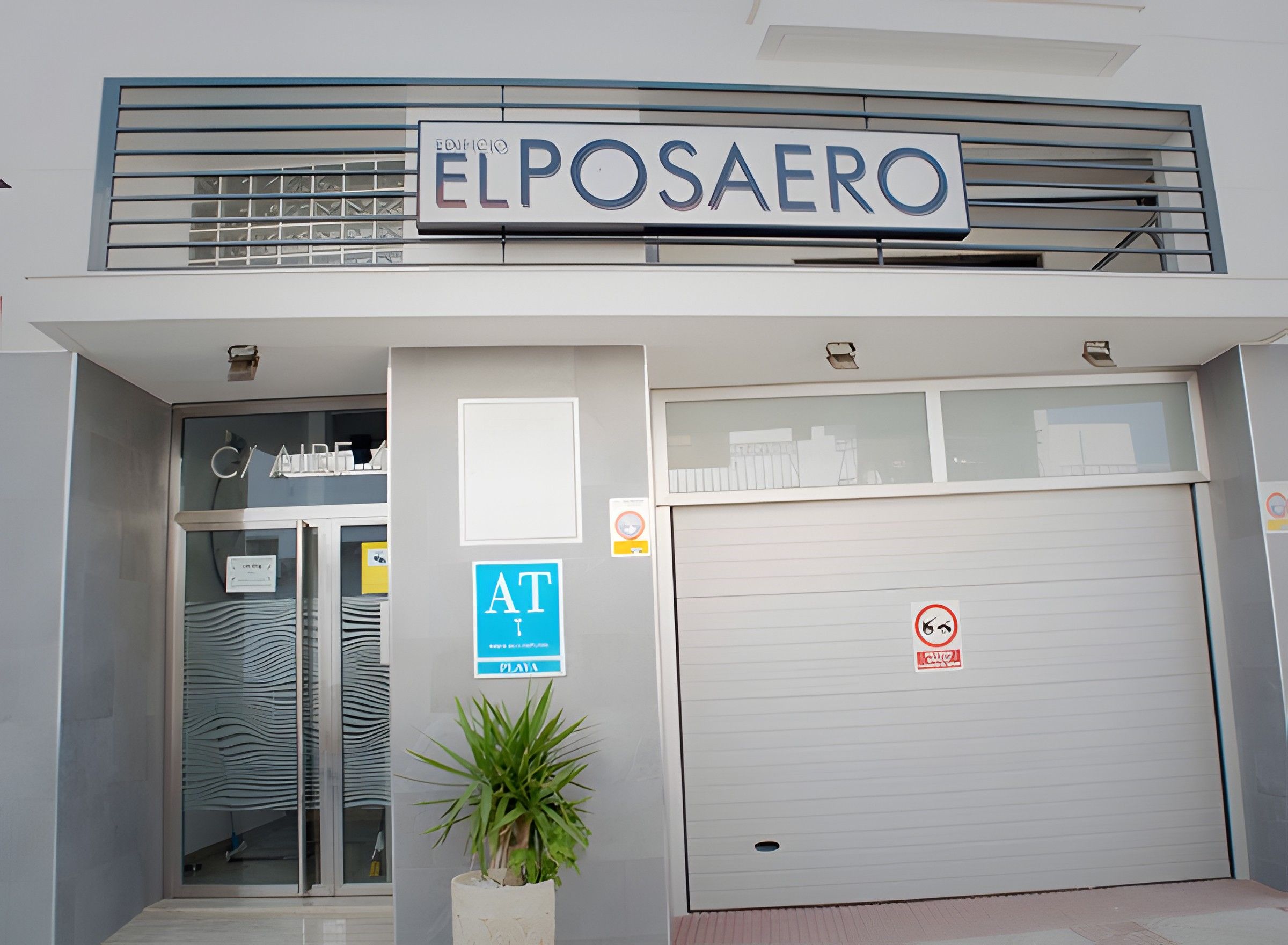 A building with a sign reading 'ELPOSAERO' above the entrance, a glass door, a blue 'AT' sign, and a no-smoking sign on the garage door.