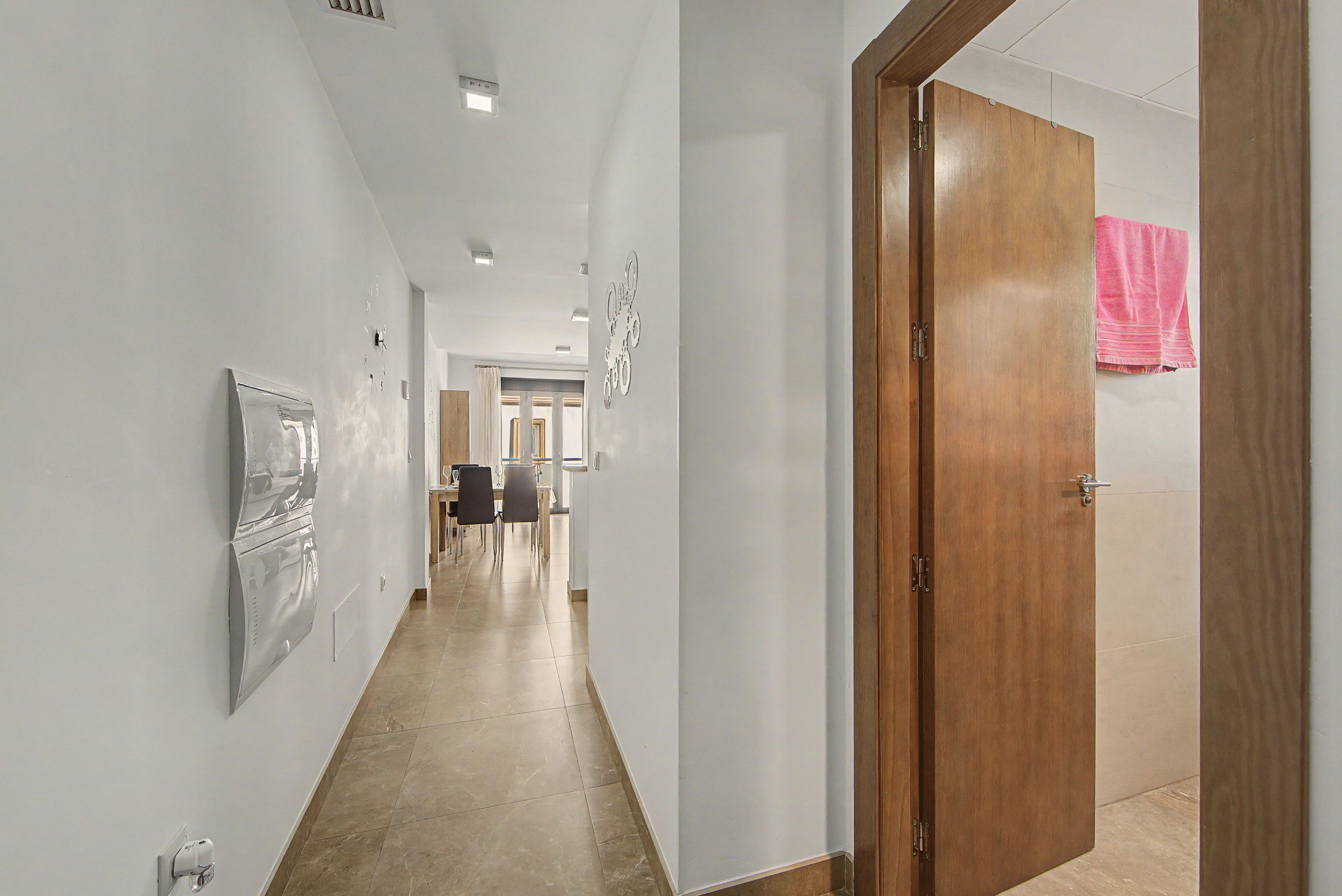 Un pasillo con una puerta de madera, una lámpara de pared y suelo de baldosas.