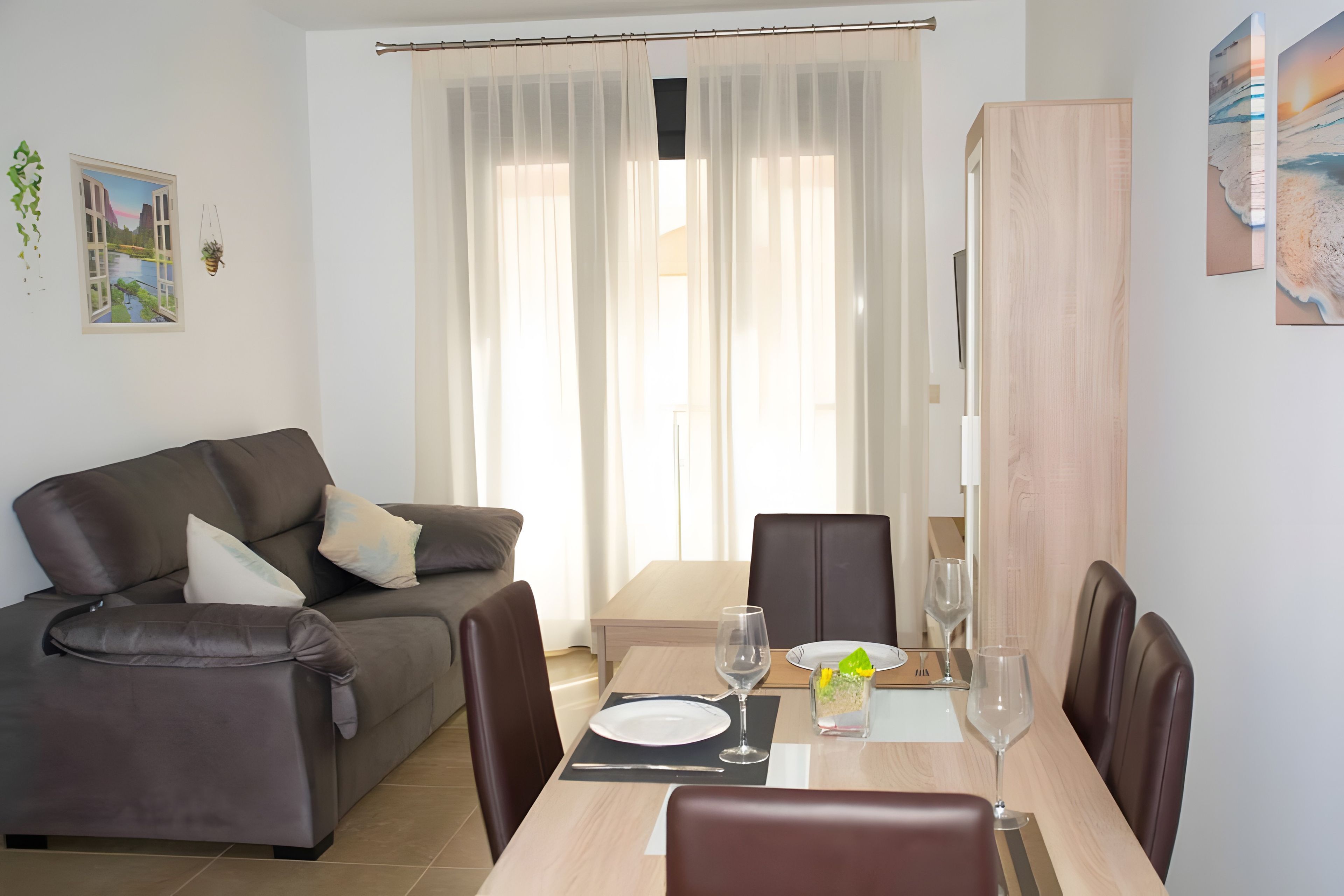 Een moderne woonkamer met een bank, een eettafel voor twee personen en een groot raam met dunne gordijnen.