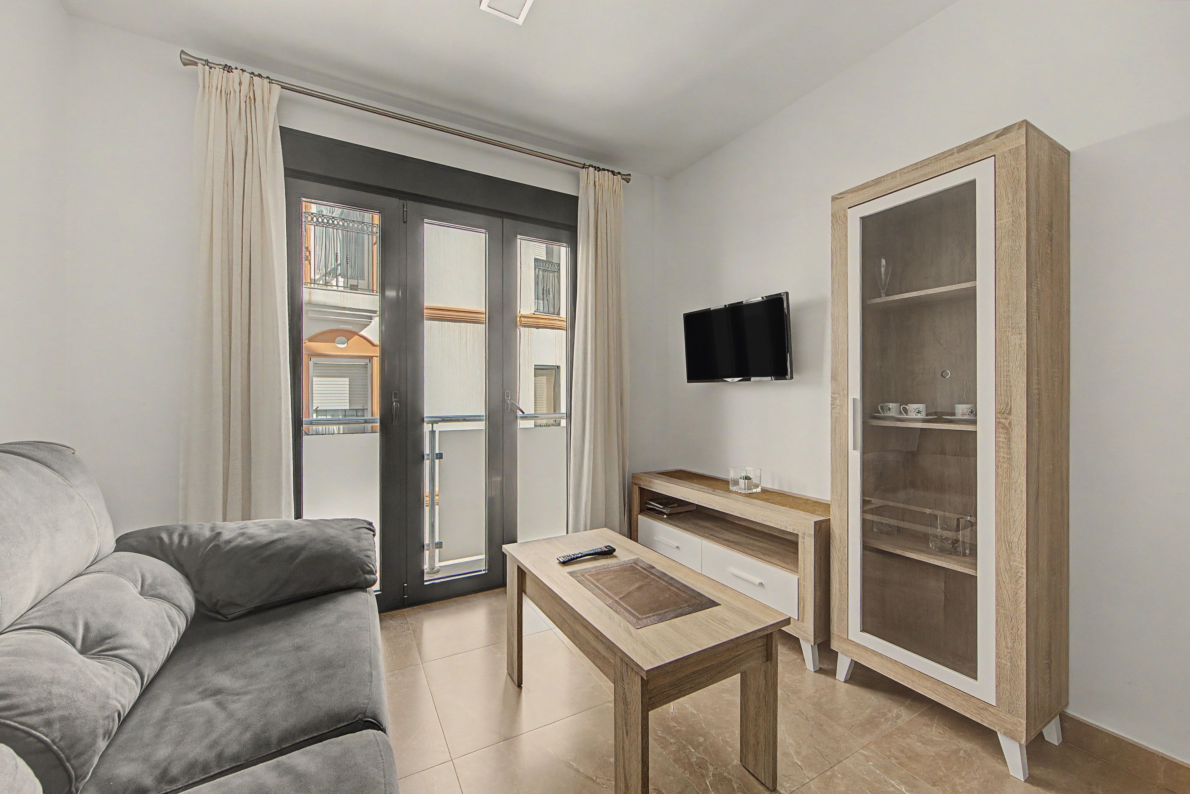 Een woonkamer met een grijze bank, een houten salontafel, een flatscreen-televisie aan de muur en een houten kast met glazen deuren.
