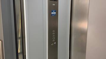An elevator interior with a metallic finish, a control panel with buttons, and a sign indicating the elevator's name.