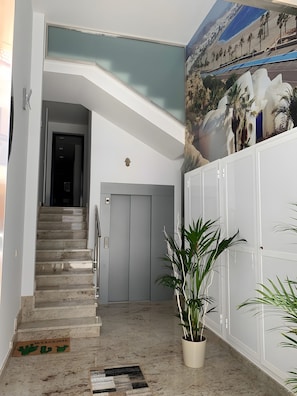 Een modern interieur met een trap naar een lift, een deurmat met cactussen en een muurschildering van een strand.