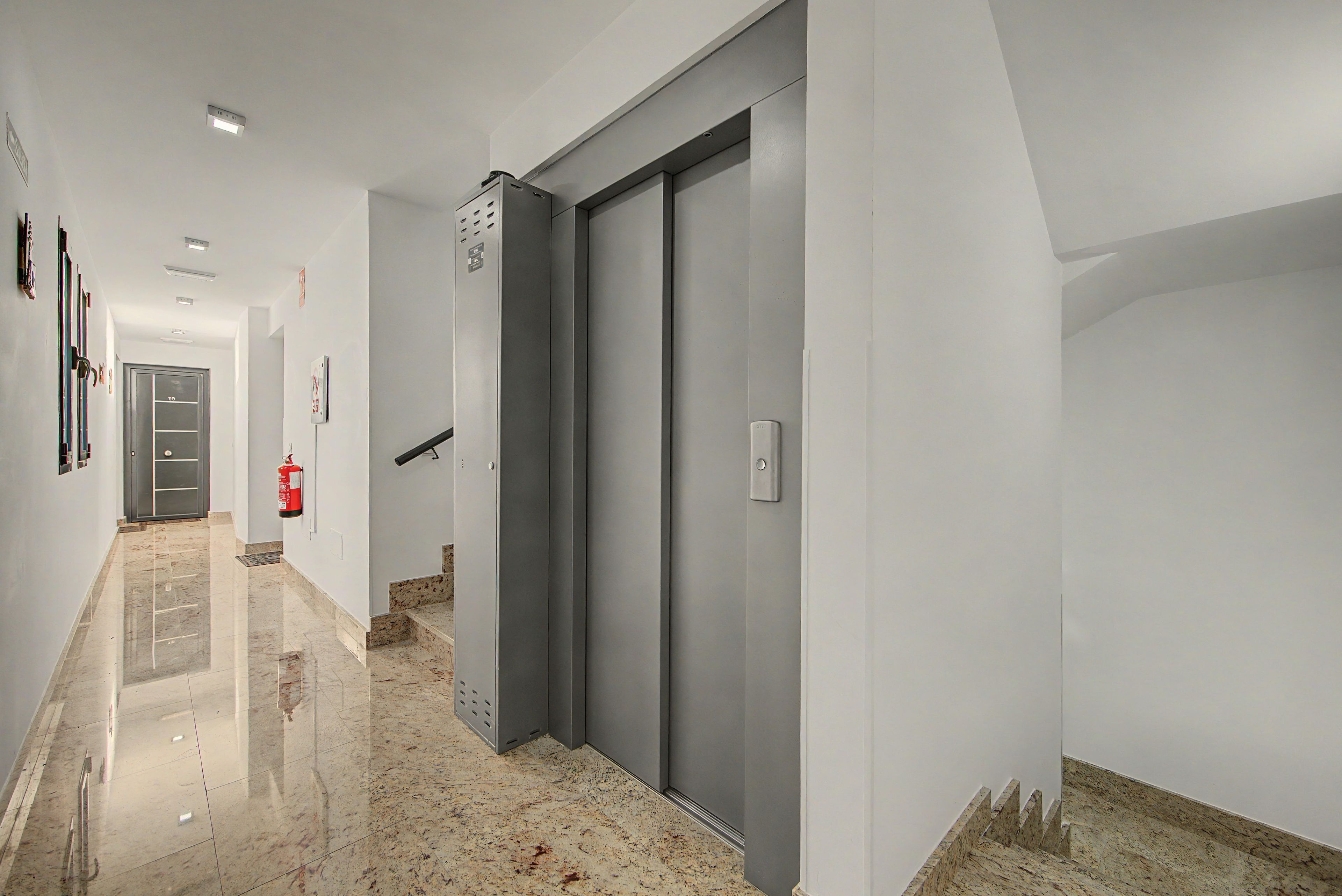 Um corredor com extintor, uma porta e luminária fixada na parede.