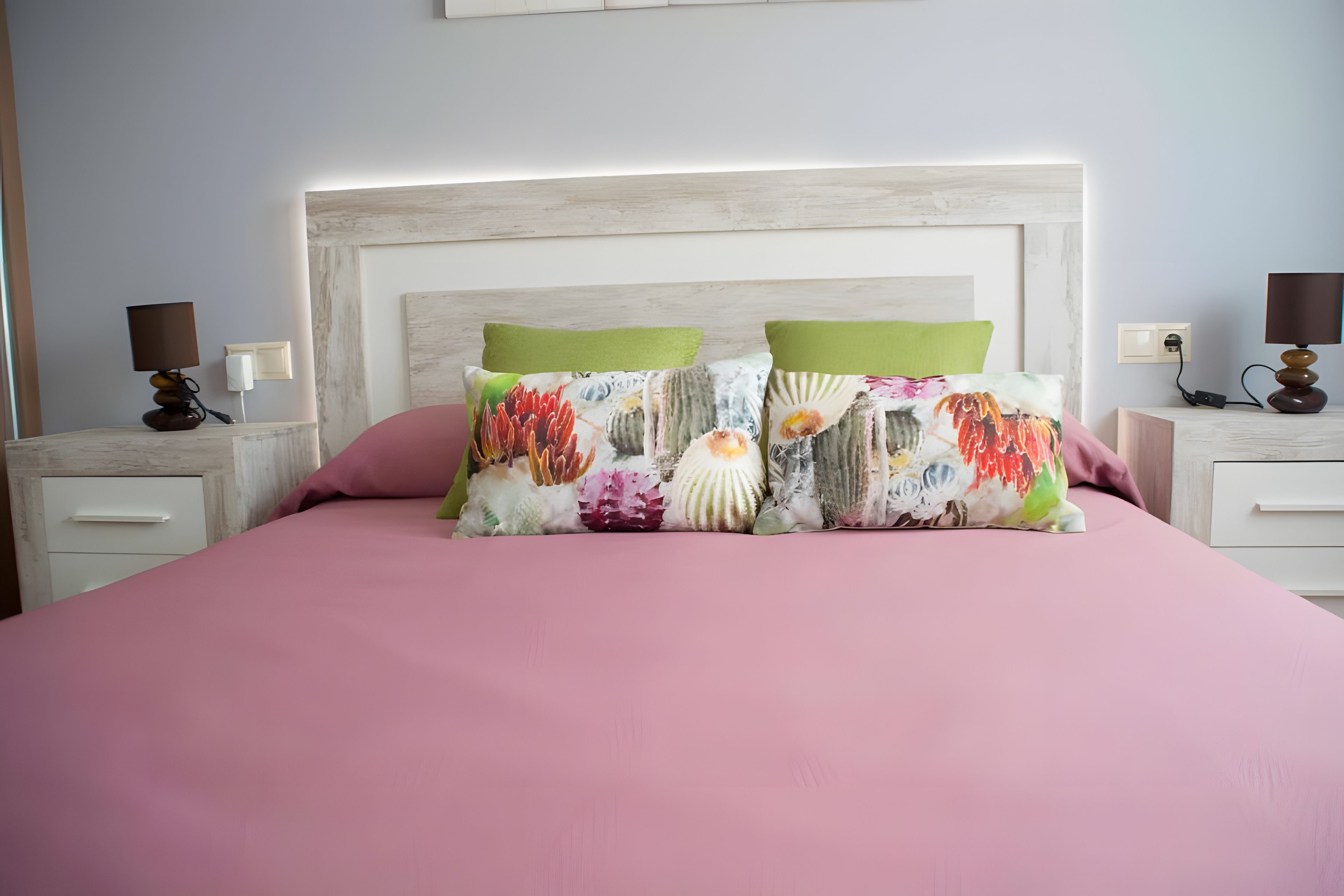 Ein Bett mit einer rosa Bettdecke, zwei Kissen mit floralen Mustern, ein weißes Kopfteil und Nachttischlampen auf Nachttischen.