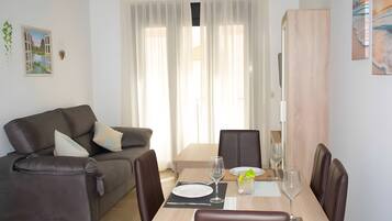 A modern living room with a sofa, a dining table set for two, and a large window with sheer curtains.