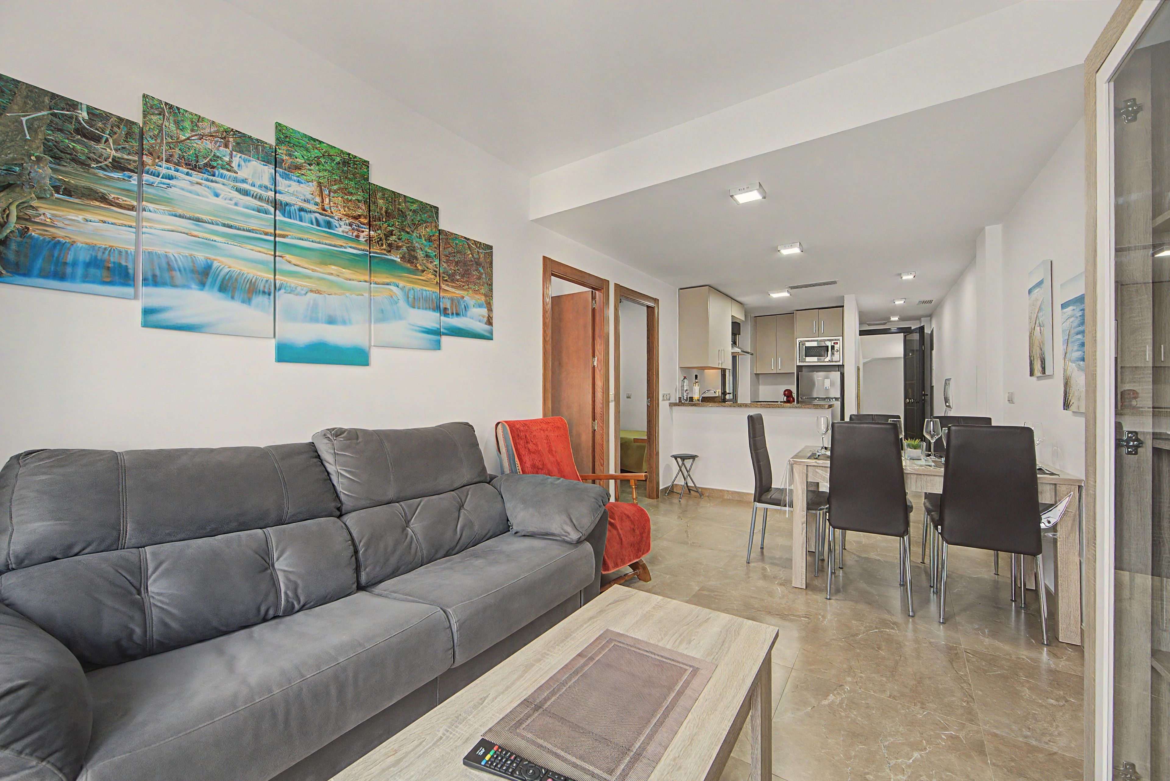Uma sala de estar moderna com um sofá cinzento, uma área de jantar com mesa e cadeiras, e uma cozinha ao fundo.