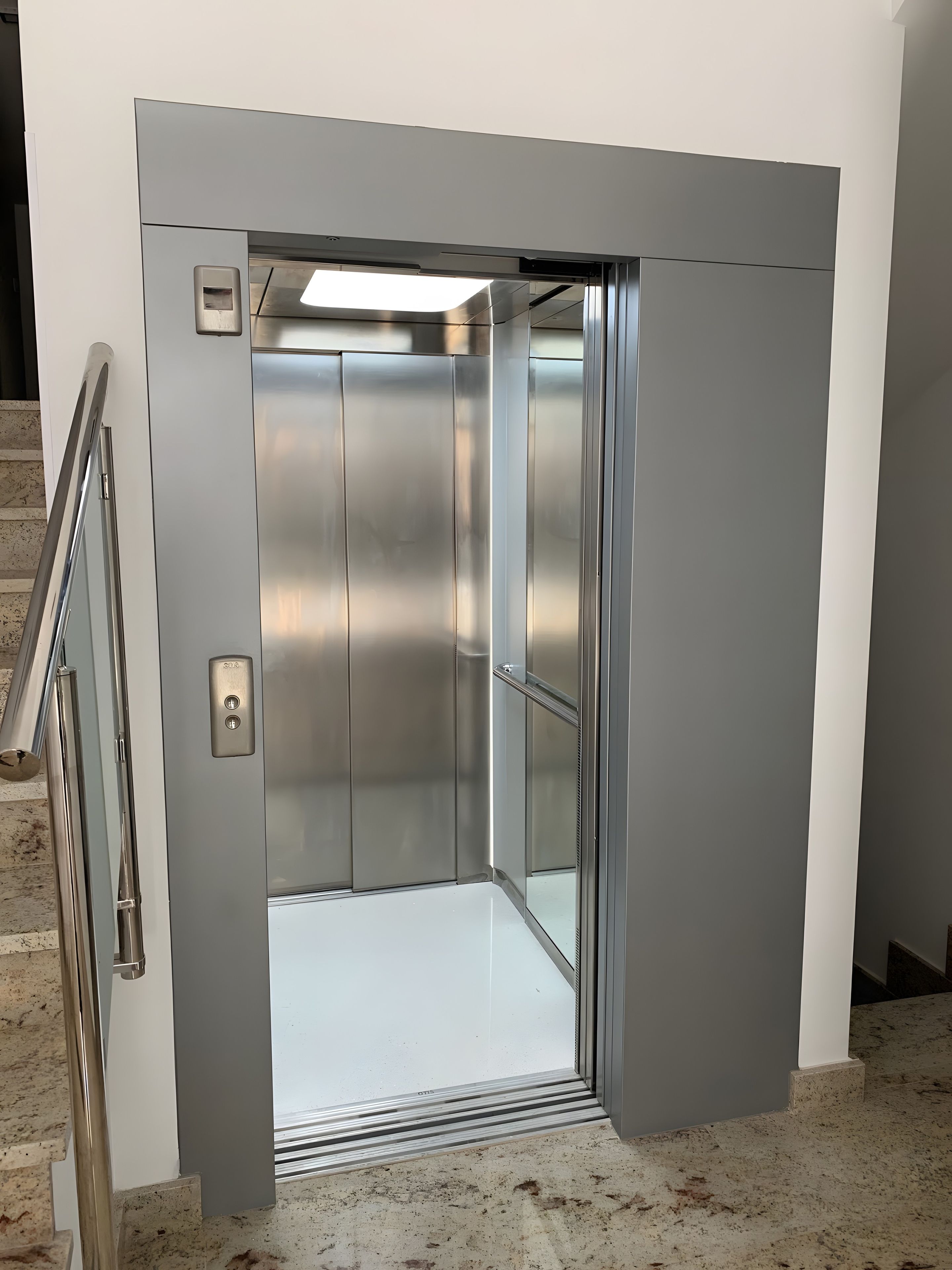A metallic elevator with a control panel, a handrail, and a glass door.