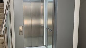 A metallic elevator with a control panel, a handrail, and a glass door.