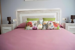 Een bed met een roze sprei, twee kussens met bloemmotieven, een wit hoofdbord en nachtlampjes op nachtkastjes.