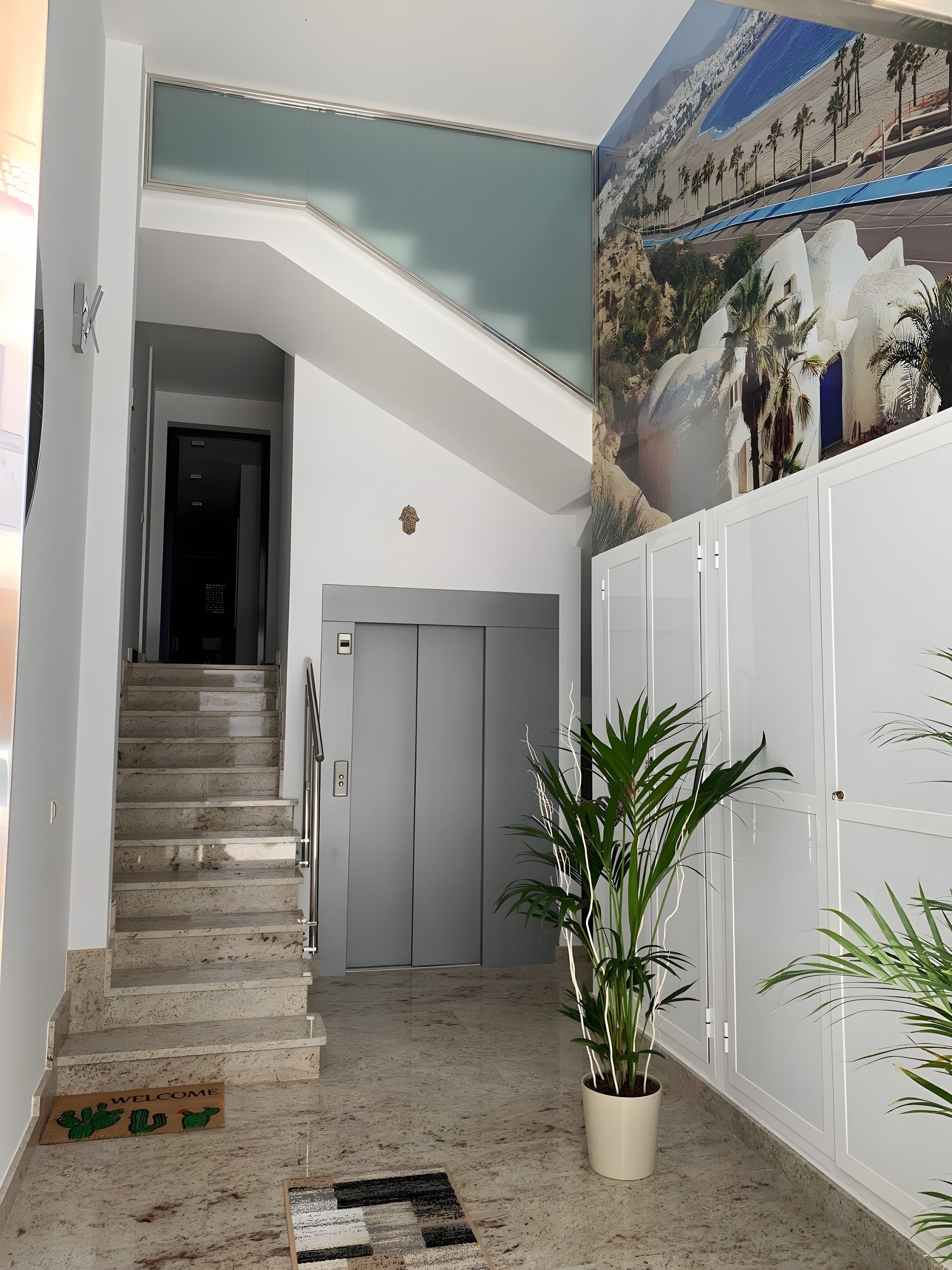 A modern interior with a staircase leading to an elevator, a welcome mat with cacti, and a mural of a beach scene.