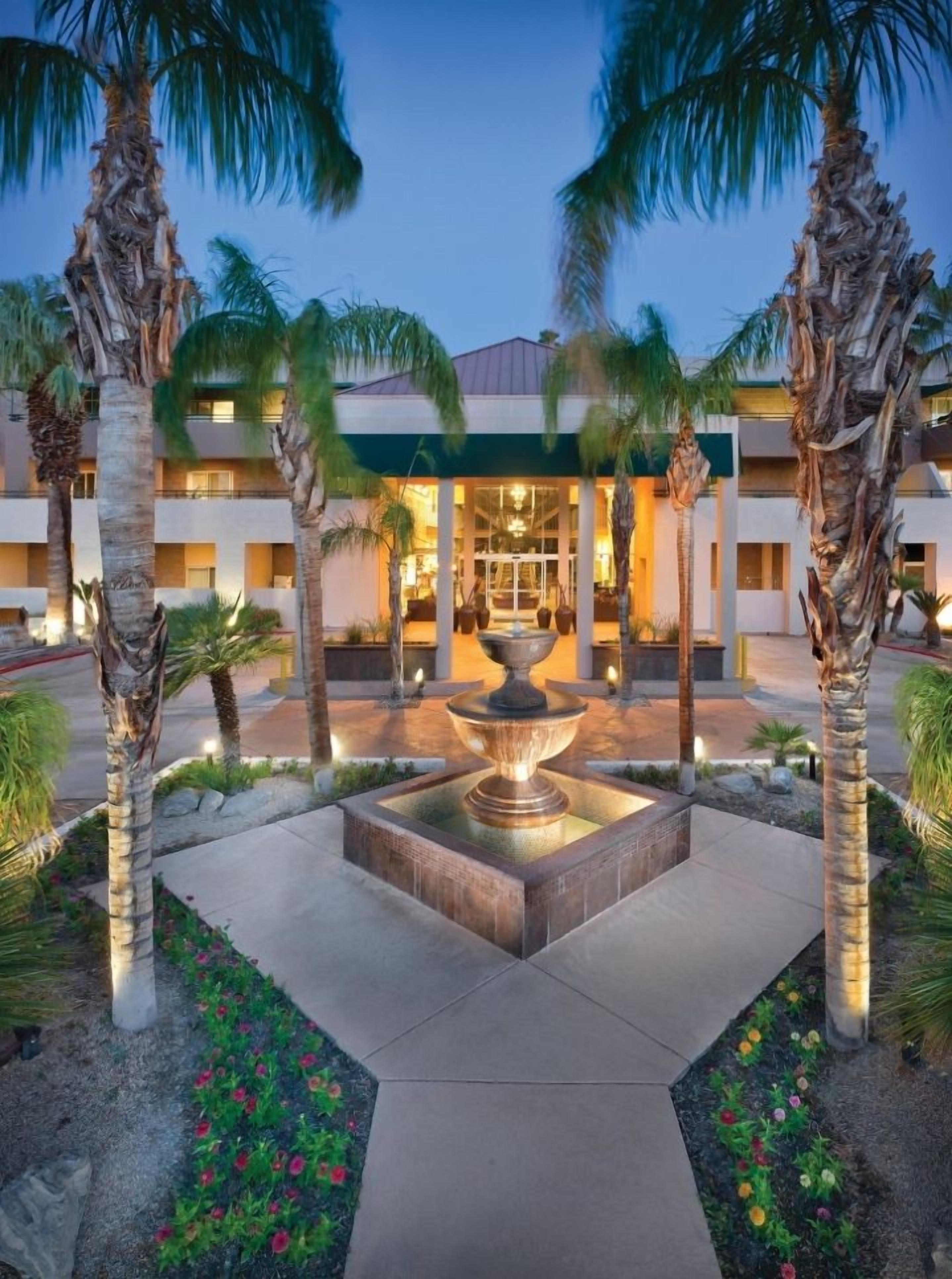 A hotel entrance with a central fountain, palm trees, and a well-lit building facade.