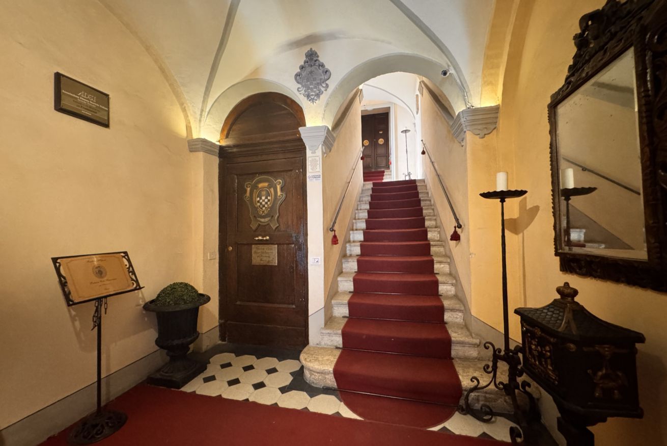 Eine prachtvolle Eingangshalle mit rotem Teppich, der zu einer Treppe führt, einer Holztür mit einer Schrifttafel und einem großen Spiegel auf der rechten Seite.