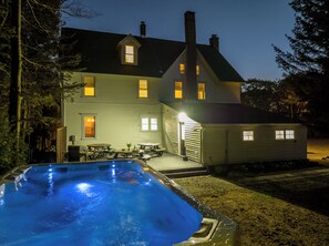 Une maison avec une piscine, une terrasse dotée de tables et de chaises, et un éclairage extérieur agréable au crépuscule.