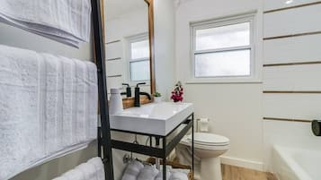 A modern bathroom with a white sink, black faucet, and a mirror. There is a toilet and a window above it.