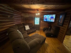 Een interieur van een blokhut met een hoekbank, een aan de muur bevestigde televisie en een lijst met een foto.