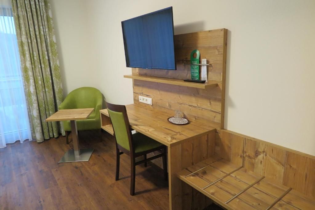 Ein kleines Zimmer mit einem Holztisch, einem grünen Stuhl, einem an der Wand montierten Fernseher und einem Regal mit einer Flasche und einem kleinen Gegenstand.