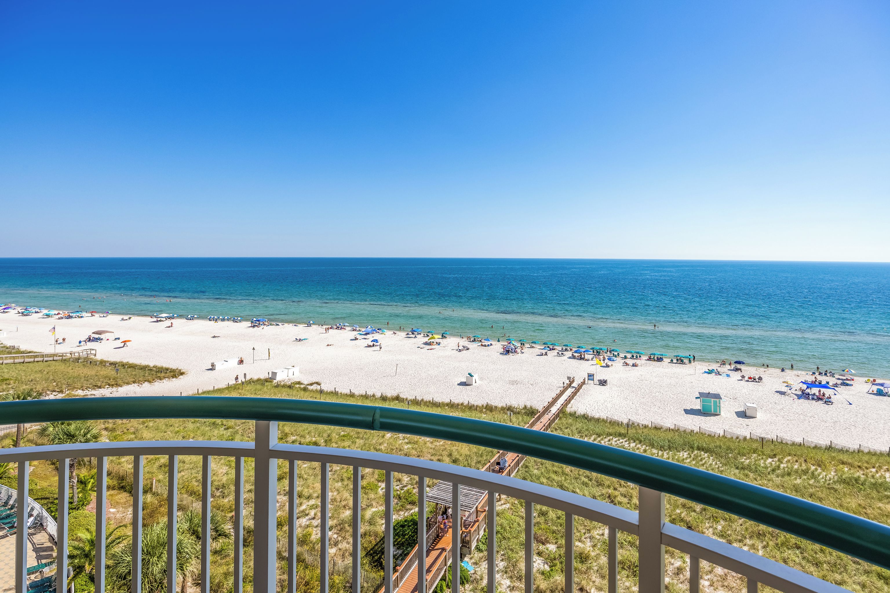 A beach view from a balcony with a clear sky, sandy beach, and a few umbrellas.