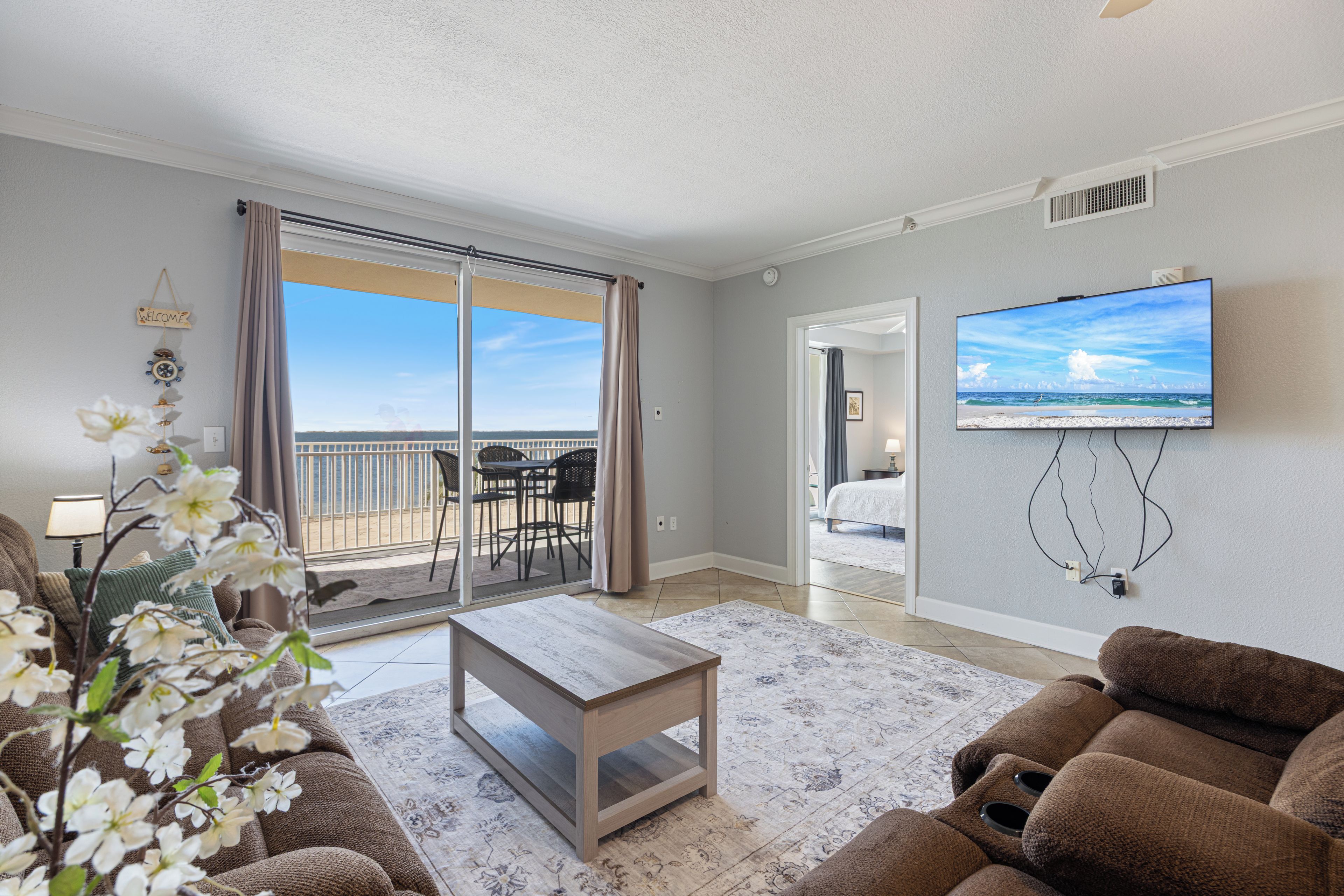 A living room with a sofa, a coffee table, a TV mounted on the wall, a balcony with a view of the ocean, and a door leading to another room.