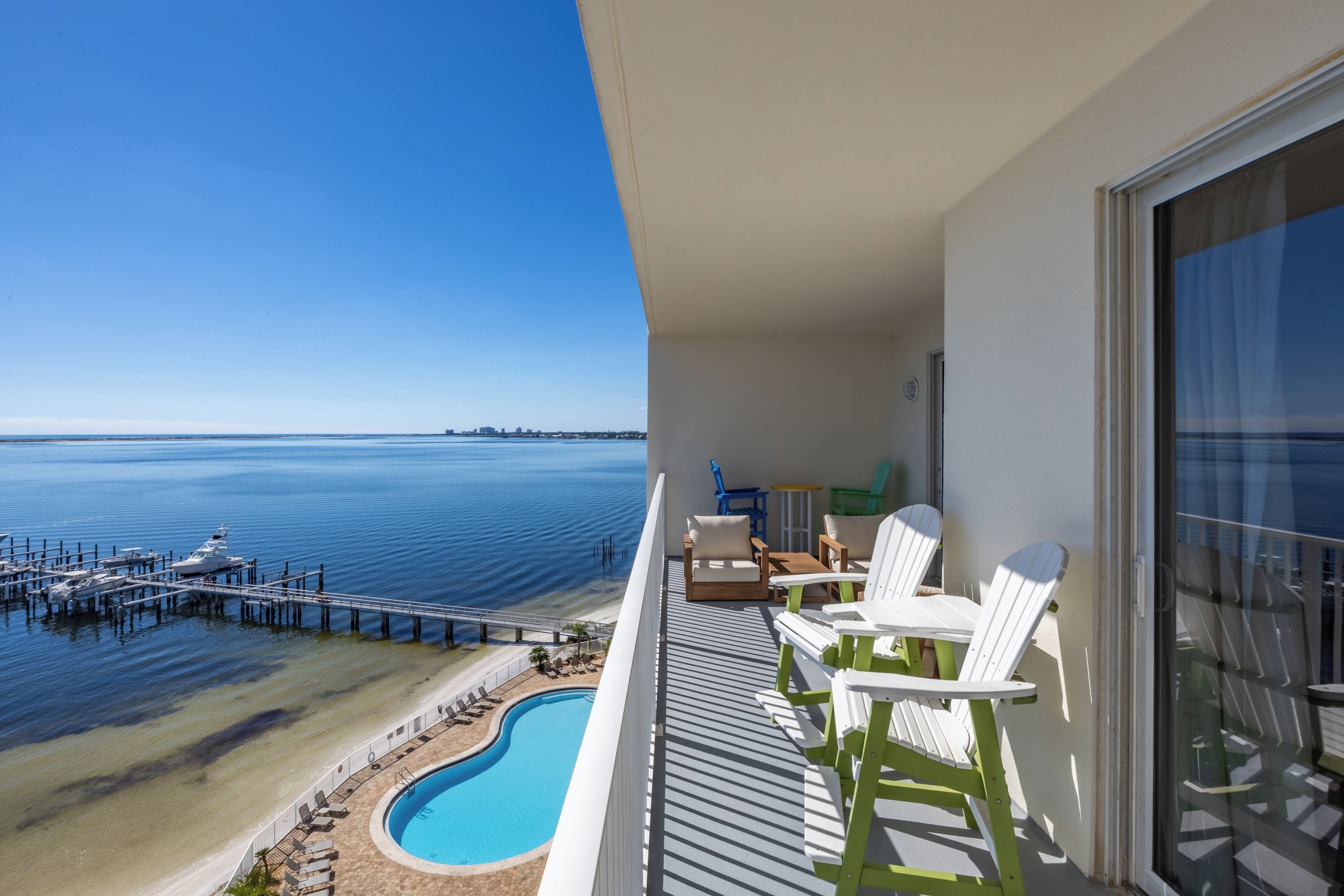 A balcony with a pool, white chairs, and a view of the ocean and pier.