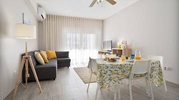 A modern living room with a sofa, a dining table with chairs, a ceiling fan, and a large window with curtains.