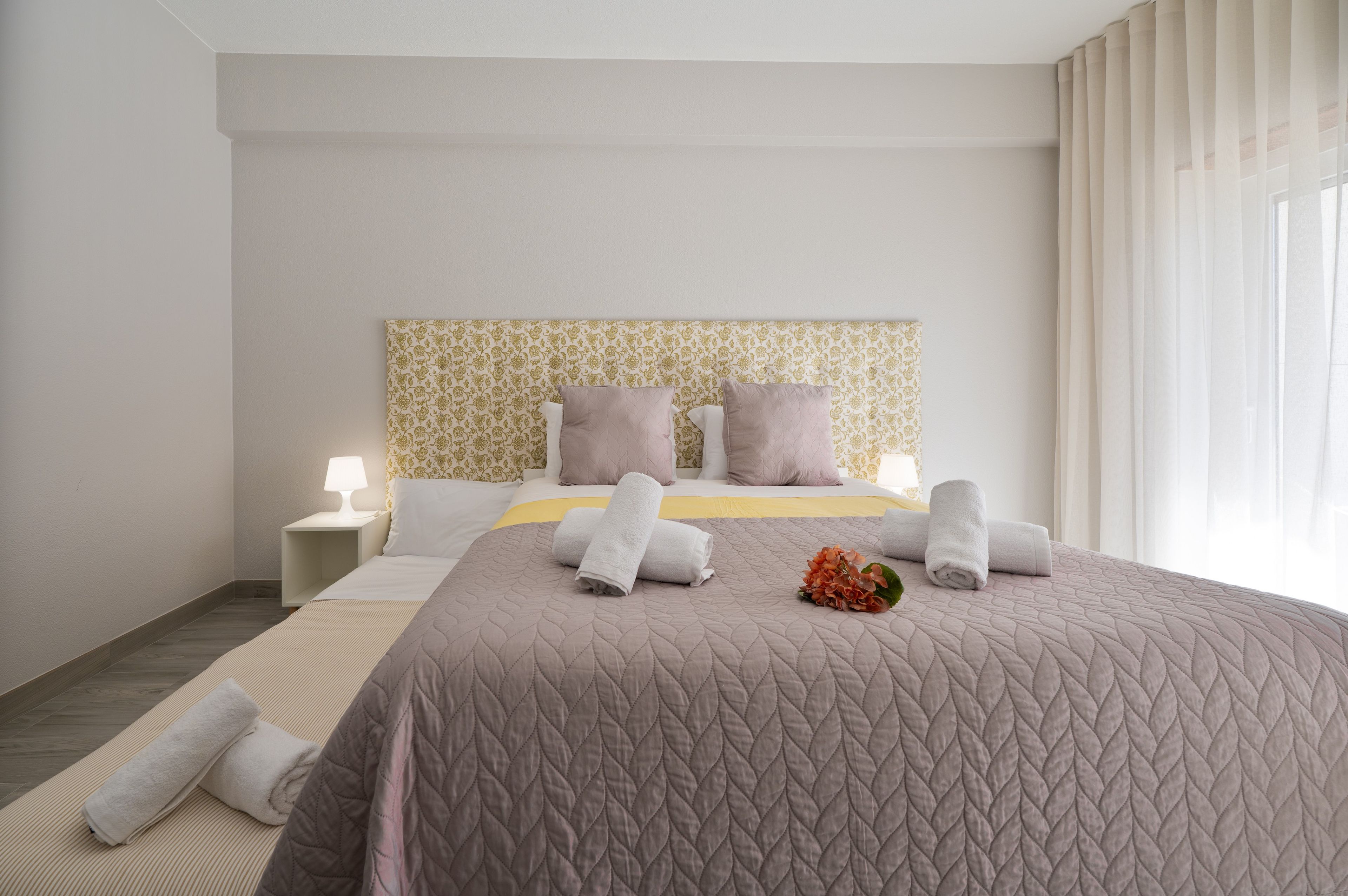 A hotel room with a bed, pillows, a bedside table with a lamp, and a window with curtains.