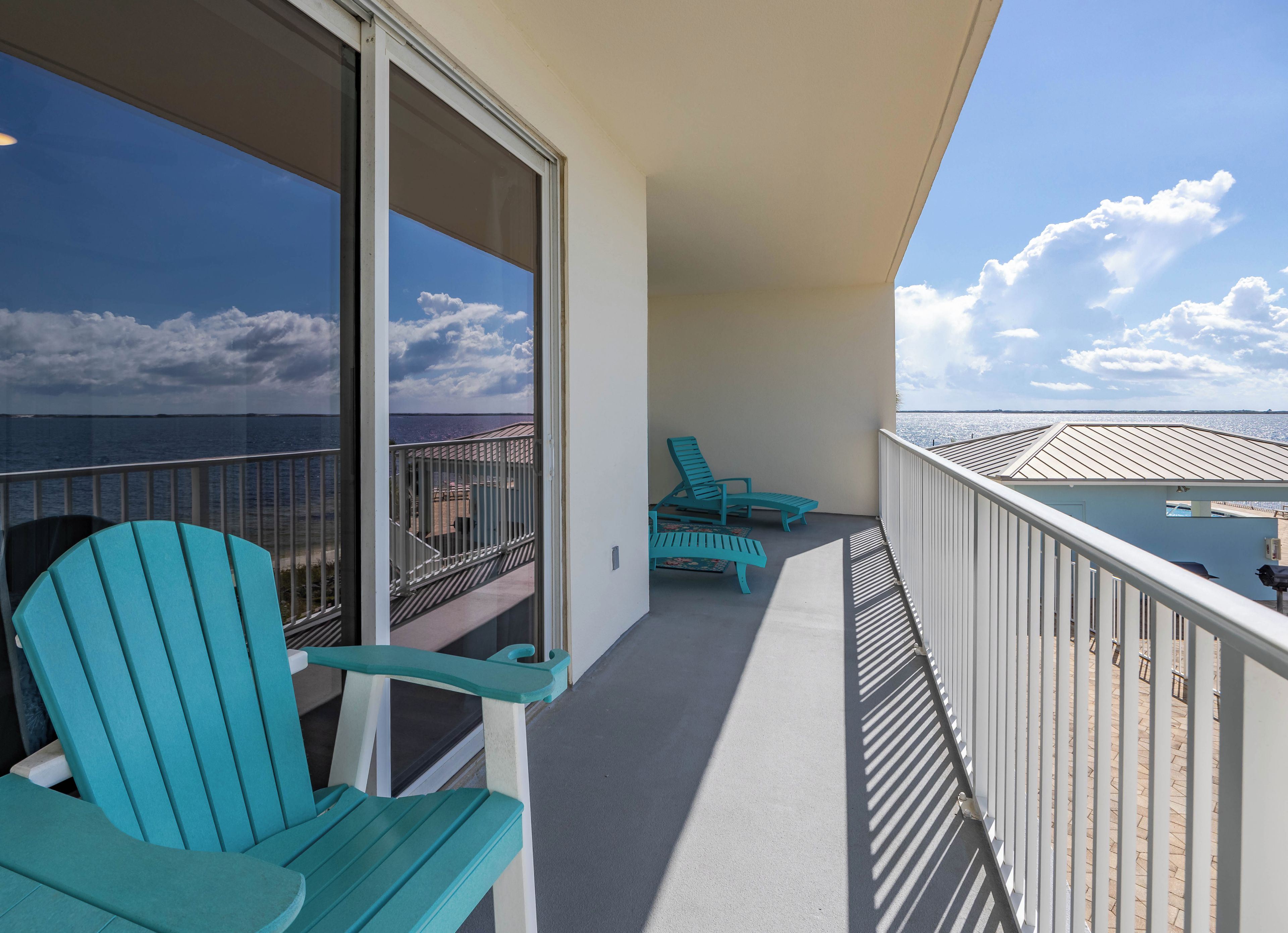 A balcony with two turquoise chairs, a glass door leading to a view of the ocean, and a railing.