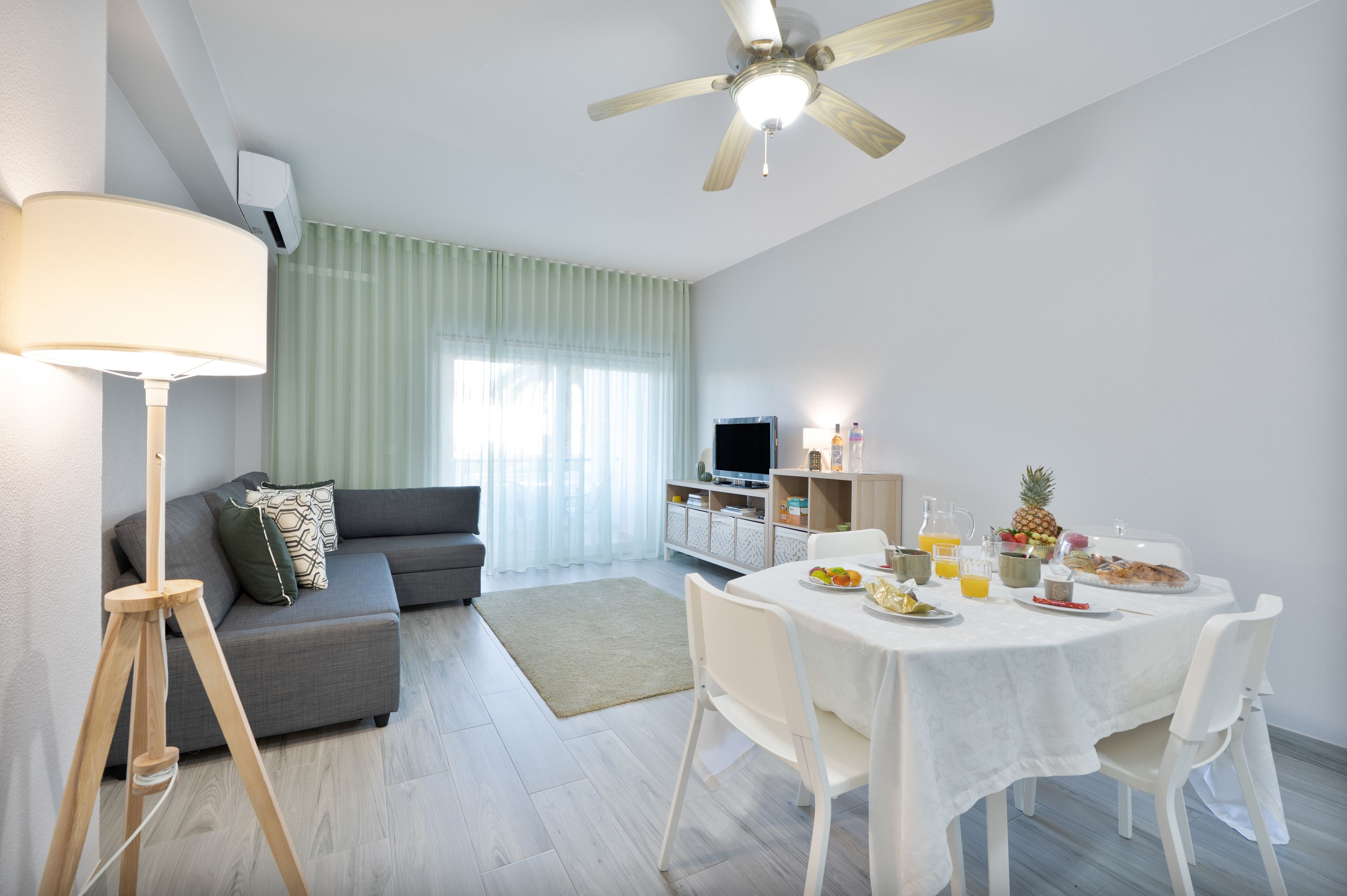 Ett modernt vardagsrum med en grå soffa, en golvlampa, ett matbord dukat för frukost och en takfläkt.