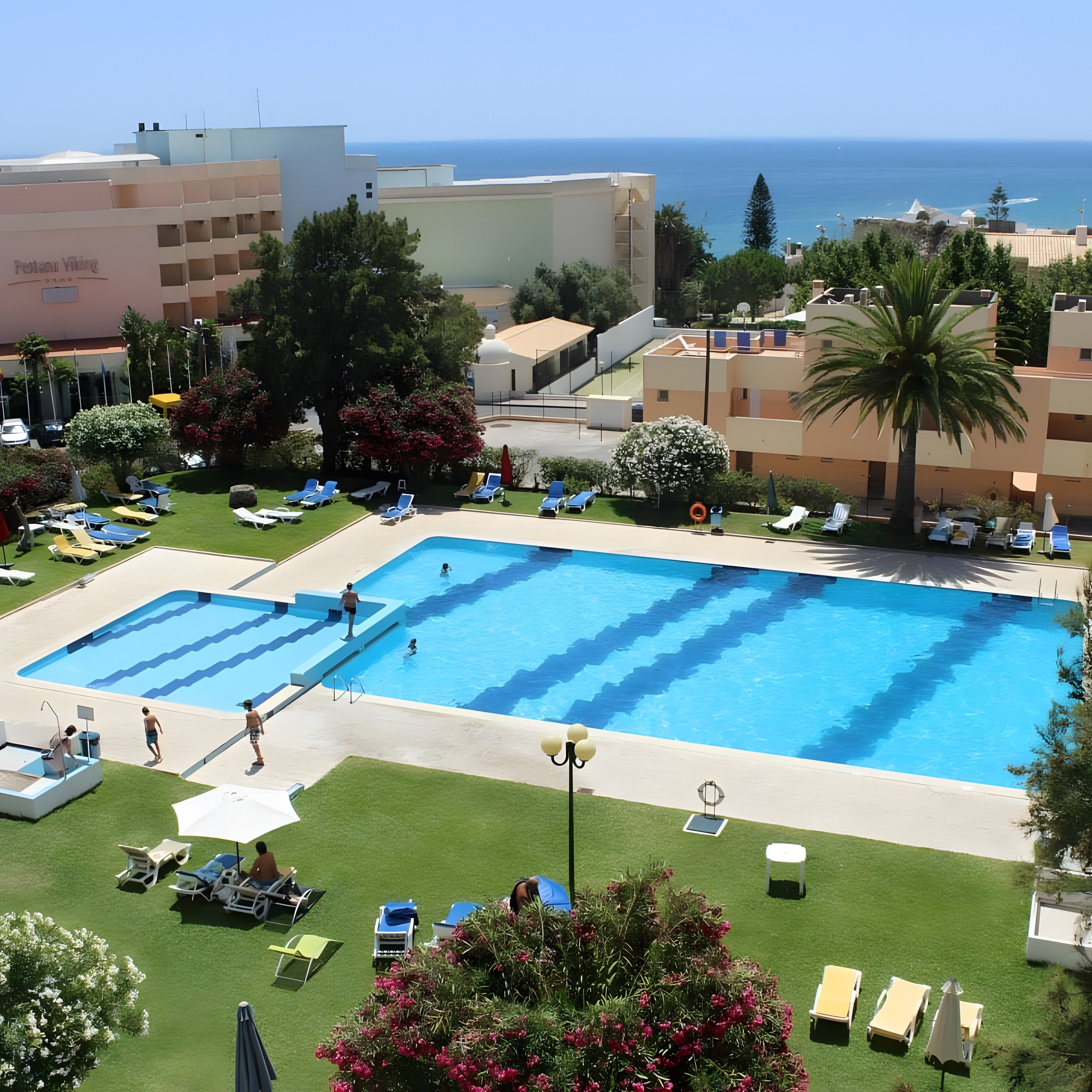 A swimming pool with multiple lanes, surrounded by lounge chairs and umbrellas, with a view of the sea and buildings in the background.