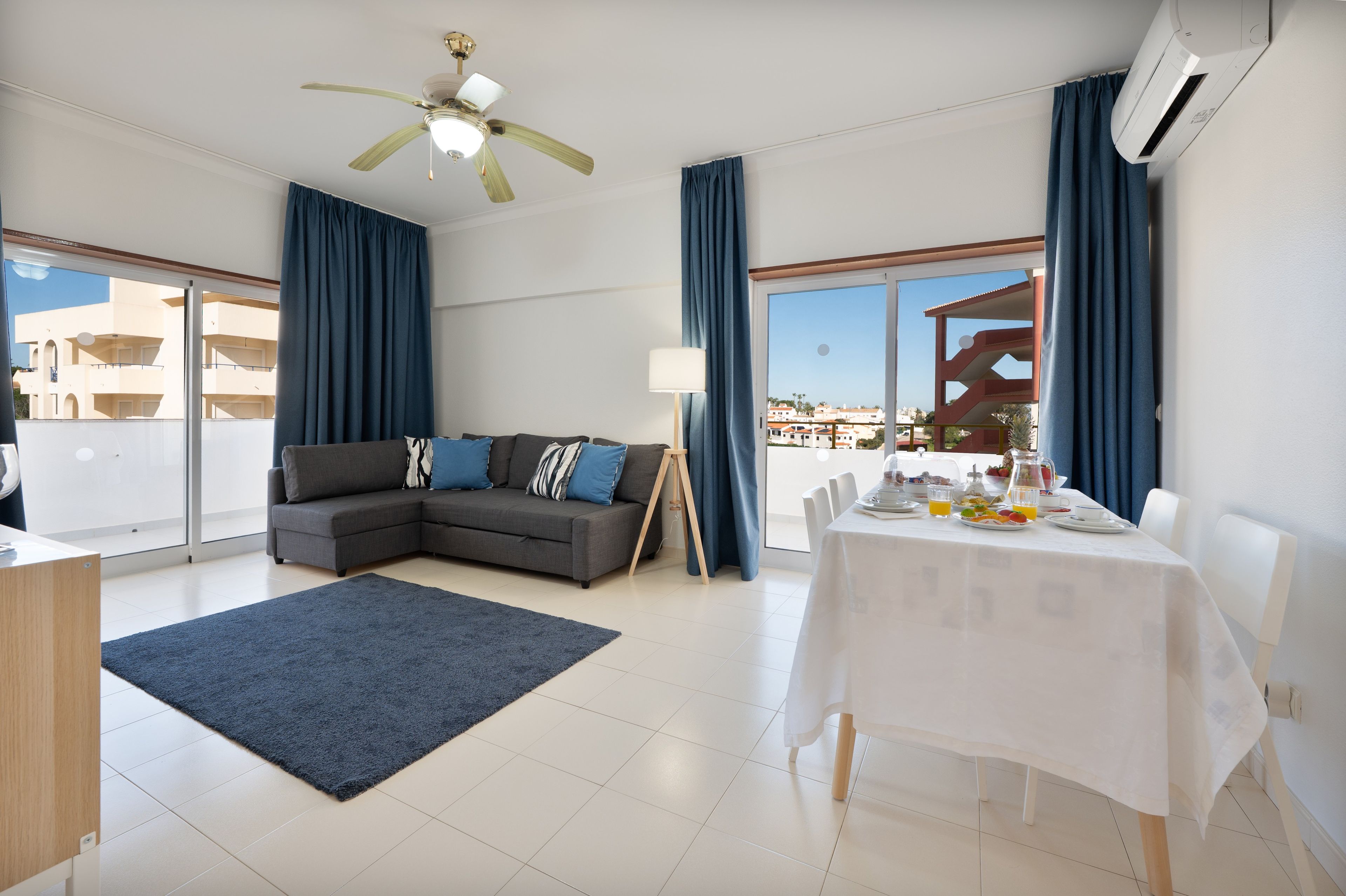 A modern living room with a sectional sofa, a dining table set for breakfast, and a large window with a view of buildings.
