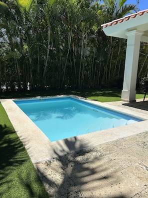 Une piscine rectangulaire dont le bord est recouvert de carreaux, entourée d’une terrasse en béton et d’une zone herbeuse.