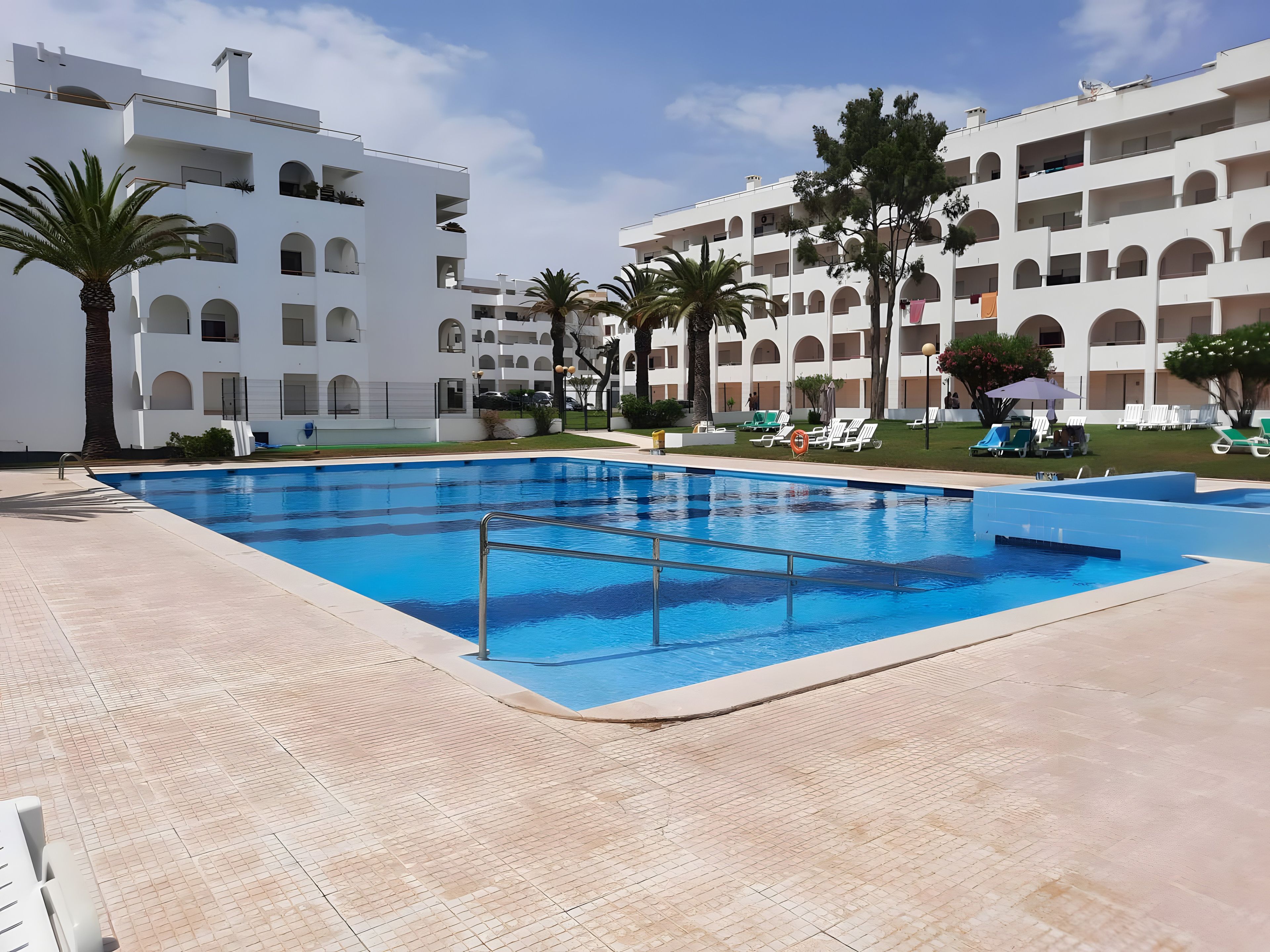 A swimming pool with a tiled deck, surrounded by white buildings with multiple balconies and palm trees.