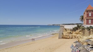 Ein Strand mit einer Steinmauer, einem Gebäude mit roter Fassade und blauem, wolkenlosem Himmel.