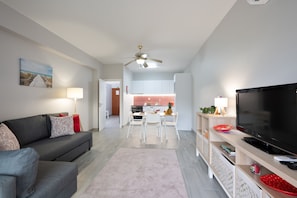 Ein modernes Wohnzimmer mit einem grauen Sofa, einem Flachbildfernseher und einem Essbereich, wobei im Hintergrund eine Küche zu sehen ist.
