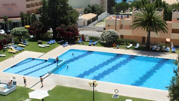 Una piscina con varias pistas, rodeada de sillas de descanso y sombrillas, con vista al mar y edificios al fondo.