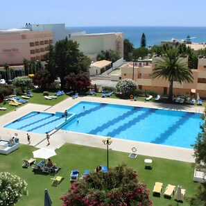 Una piscina con varias pistas, rodeada de sillas de descanso y sombrillas, con vista al mar y edificios al fondo.