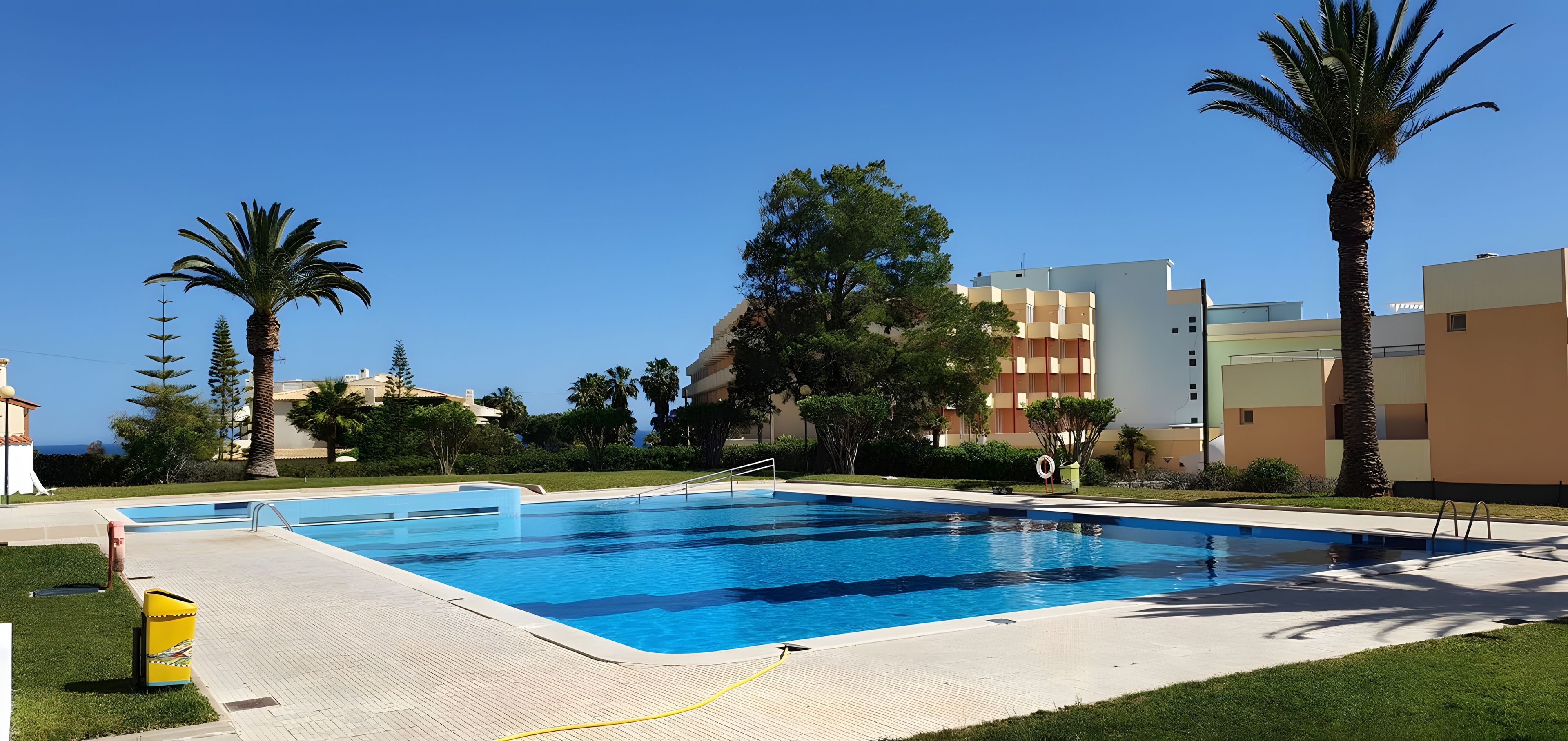 A swimming pool with a clear blue water surface, surrounded by a paved deck and a grassy area.